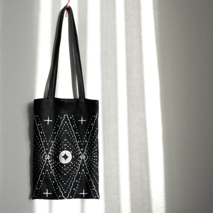 Black denim tote bag with white star-pattern Sashiko embroidery hanging against a sunlit wall.