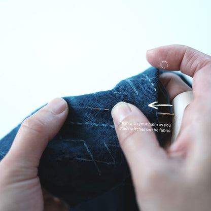 Pushing a needle with the leather palm thimble while stacking stitches on indigo fabric.